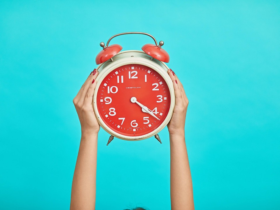 hands holding up a red alarm clock on a bright blue background