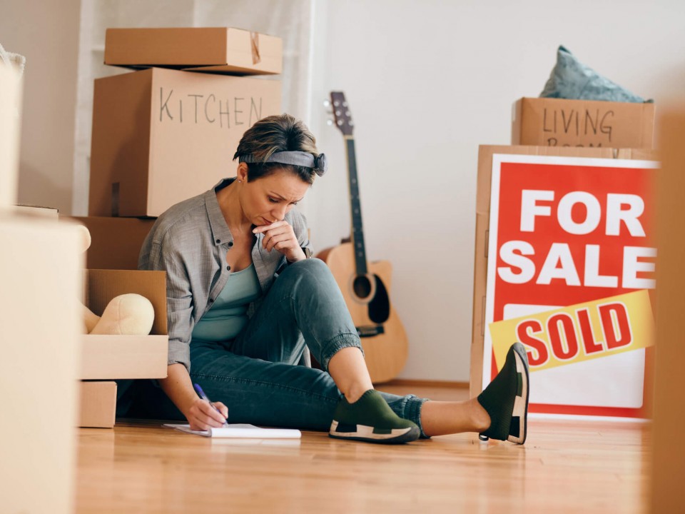 woman looking concerned sitting on the floor making notes surrounded by packing boxes and a 'sold' house sign